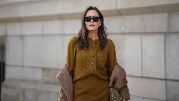 Laura Noltemeyer is seen wearing a a long camel-colored brushed wool coat with a wrap silhouette and integrated belt by Max Mara; a mustard-colored knit cardigan with a brushed cable-knit pattern and front button fastening by Sézane; a small quilted tweed flap bag in a khaki Color with woven fabric material with a gold chain strap and CC clasp by Chanel; gold diamond rings by Ariane Ernst; gold statement earrings with a ruffled surface and a green stone element by Mango; black cat eye sunglasses with an acetate frame by Yves Saint Laurent; her brown hair is styled in loose waves with a center part on November 13, 2025 in Hamburg, Germany.