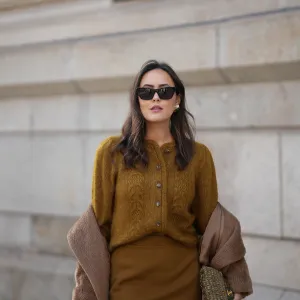 Laura Noltemeyer is seen wearing a a long camel-colored brushed wool coat with a wrap silhouette and integrated belt by Max Mara; a mustard-colored knit cardigan with a brushed cable-knit pattern and front button fastening by Sézane; a small quilted tweed flap bag in a khaki Color with woven fabric material with a gold chain strap and CC clasp by Chanel; gold diamond rings by Ariane Ernst; gold statement earrings with a ruffled surface and a green stone element by Mango; black cat eye sunglasses with an acetate frame by Yves Saint Laurent; her brown hair is styled in loose waves with a center part on November 13, 2025 in Hamburg, Germany.