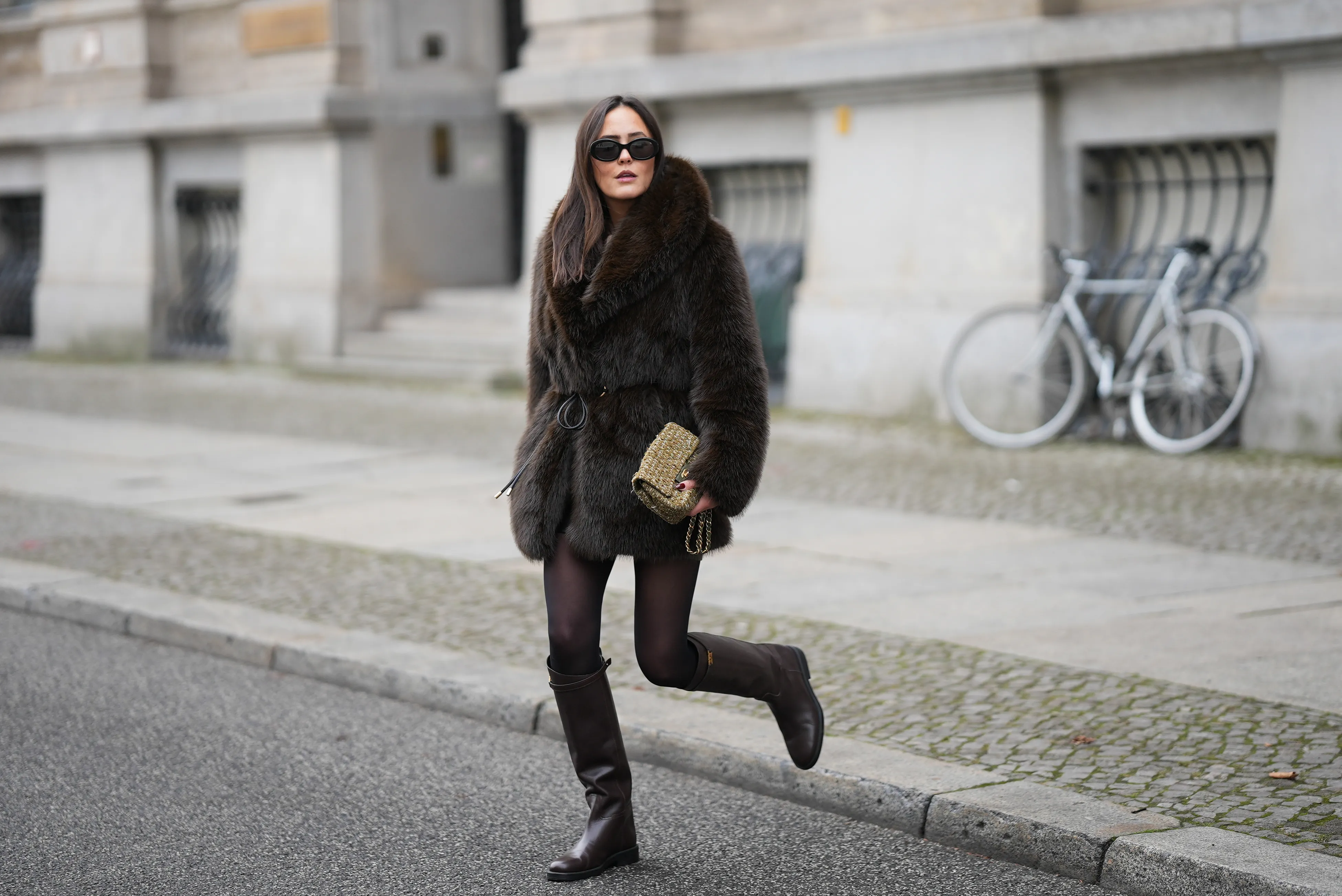 Laura Noltemeyer is seen wearing a dark-brown faux-fur coat with a plush, heavyweight texture, wide notch lapels and a straight hip-length silhouette with a hook and eye belt closing from Zara; a cream knit sweater with a soft ribbed crew neckline from S&eacute;zane; semi-sheer black tights; knee-high dark brown leather riding boots with a straight shaft, gold buckle detail and a flat sole from Massimo Dutti; a small quillted and woven flap bag in a khaki green tweed color featuring the signature CC turn-lock and a gold hardware chain strap from Chanel; black oval sunglasses with a glossy acetate frame and dark lenses from Celine; chunky sculptural gold earrings from Zara; her dark brown hair is worn straight with a center part on November 25, 2025 in Berlin, Germany.