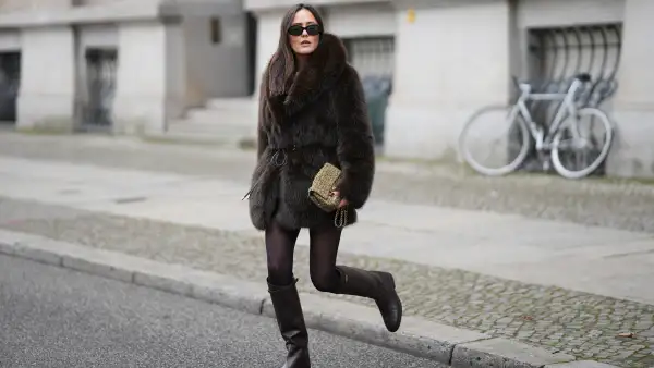 Laura Noltemeyer is seen wearing a dark-brown faux-fur coat with a plush, heavyweight texture, wide notch lapels and a straight hip-length silhouette with a hook and eye belt closing from Zara; a cream knit sweater with a soft ribbed crew neckline from Sézane; semi-sheer black tights; knee-high dark brown leather riding boots with a straight shaft, gold buckle detail and a flat sole from Massimo Dutti; a small quillted and woven flap bag in a khaki green tweed color featuring the signature CC turn-lock and a gold hardware chain strap from Chanel; black oval sunglasses with a glossy acetate frame and dark lenses from Celine; chunky sculptural gold earrings from Zara; her dark brown hair is worn straight with a center part on November 25, 2025 in Berlin, Germany.