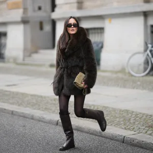 Laura Noltemeyer is seen wearing a dark-brown faux-fur coat with a plush, heavyweight texture, wide notch lapels and a straight hip-length silhouette with a hook and eye belt closing from Zara; a cream knit sweater with a soft ribbed crew neckline from Sézane; semi-sheer black tights; knee-high dark brown leather riding boots with a straight shaft, gold buckle detail and a flat sole from Massimo Dutti; a small quillted and woven flap bag in a khaki green tweed color featuring the signature CC turn-lock and a gold hardware chain strap from Chanel; black oval sunglasses with a glossy acetate frame and dark lenses from Celine; chunky sculptural gold earrings from Zara; her dark brown hair is worn straight with a center part on November 25, 2025 in Berlin, Germany.