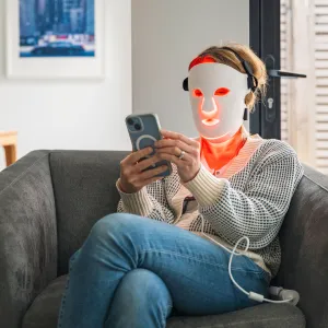 A mature woman uses her red LED face mask (red light therapy) whilst relaxing at home in an armchair. She uses her smartphone - stock photo