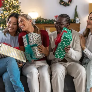 A diverse group of friends sits on a couch, smiling as they unwrap colorful gifts during a Christmas celebration at home. A young woman is taking selfie