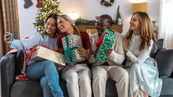 A diverse group of friends sits on a couch, smiling as they unwrap colorful gifts during a Christmas celebration at home. A young woman is taking selfie