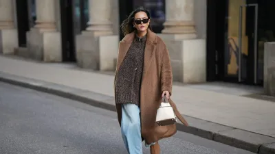 HAMBURG, GERMANY - NOVEMBER 13: Laura Noltemeyer is seen wearing a long camel-colored brushed wool wrap coat with a belt and wide lapels by Max Mara; a chunky zip-front sweater cardigan in a mélange brown wool blend with a textured, hand-knit look by by Aylin Koenig; light blue wide-leg jeans in a soft washed denim with a relaxed fit and front tag detail by Closed; chestnut colored suede slippers with plush lining called „Tasman“ by UGG; a structured bucket-shaped handbag in an off-white linen shade with taupe leather handles and lining details called „Picotin Cargo 18 Toile Goeland“ by Hermès; black cat eye sunglasses with an acetate frame by Yves Saint Laurent; gold statement earrings with a ruffled surface and a green stone element by Mango ; her brown hair is worn loose in soft waves with a center part on November 13, 2025 in Hamburg, Germany. (Photo by Moritz Scholz/Getty Images)
