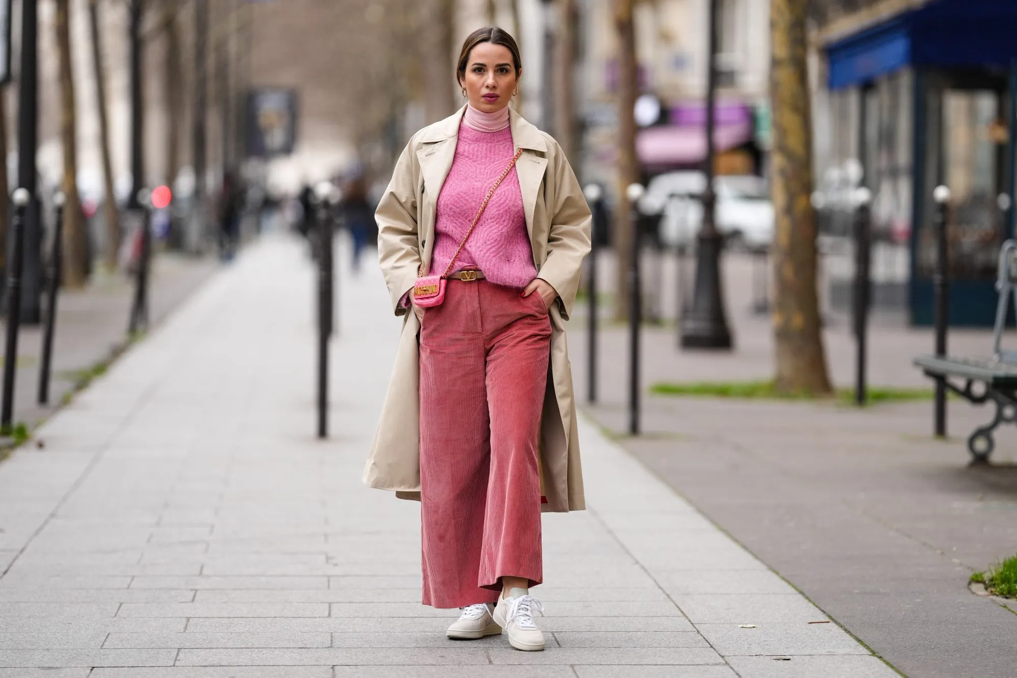 PARIS, FRANCE - FEBRUARY 04: Maria Rosaria Rizzo wears gold large earrings, a pale pink ribbed wool turtleneck pullover, a pink wool braided pattern pullover from Imprevu Belgium, a beige long trench coat from Max Mara, a pink shiny leather crossbody bag from Moschino, a beige shiny leather belt from Valentino, red ribbed velvet large pants from Max Mara, beige / pale orange and purple sneakers from Veja, during a street style fashion photo session, on February 04, 2022 in Paris, France. (Photo by Edward Berthelot/Getty Images)