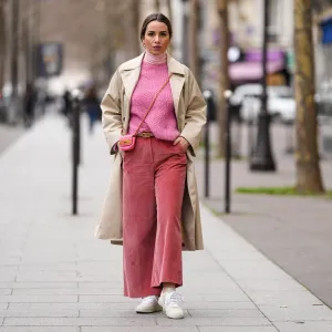 PARIS, FRANCE - FEBRUARY 04: Maria Rosaria Rizzo wears gold large earrings, a pale pink ribbed wool turtleneck pullover, a pink wool braided pattern pullover from Imprevu Belgium, a beige long trench coat from Max Mara, a pink shiny leather crossbody bag from Moschino, a beige shiny leather belt from Valentino, red ribbed velvet large pants from Max Mara, beige / pale orange and purple sneakers from Veja, during a street style fashion photo session, on February 04, 2022 in Paris, France. (Photo by Edward Berthelot/Getty Images)