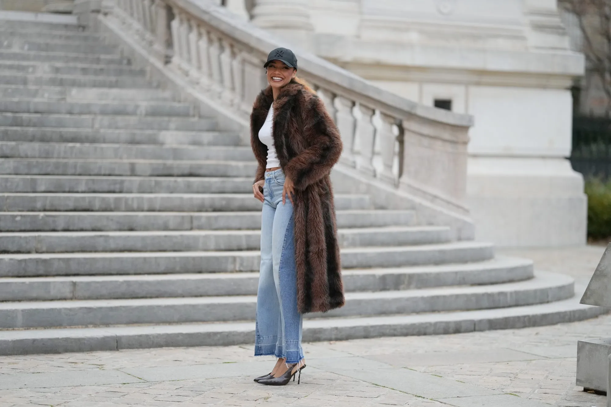 PARIS, FRANCE - DECEMBER 11: Alicia Aylies wears hair pulled back into a sleek low ponytail, natural skin with defined brows and lipstick, a black baseball NY cap, a white fitted cropped top by Celine, a long brown faux fur coat with a straight cut and button closure, light blue high waist flared wide leg jeans with a raw hem, and dark pointed toe high heel pumps shoes, during a street style fashion photo session, on December 11, 2025 in Paris, France. (Photo by Edward Berthelot/Getty Images)
