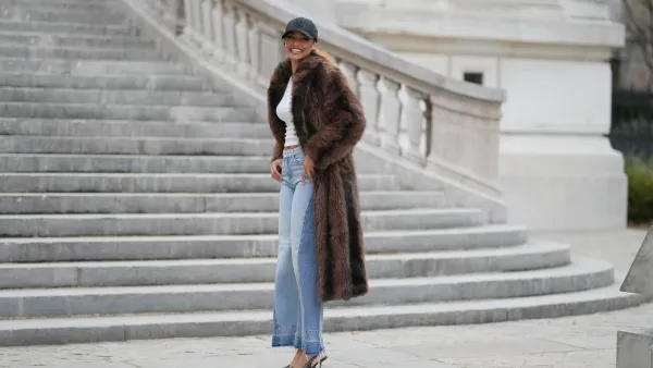 PARIS, FRANCE - DECEMBER 11: Alicia Aylies wears hair pulled back into a sleek low ponytail, natural skin with defined brows and lipstick, a black baseball NY cap, a white fitted cropped top by Celine, a long brown faux fur coat with a straight cut and button closure, light blue high waist flared wide leg jeans with a raw hem, and dark pointed toe high heel pumps shoes, during a street style fashion photo session, on December 11, 2025 in Paris, France. (Photo by Edward Berthelot/Getty Images)
