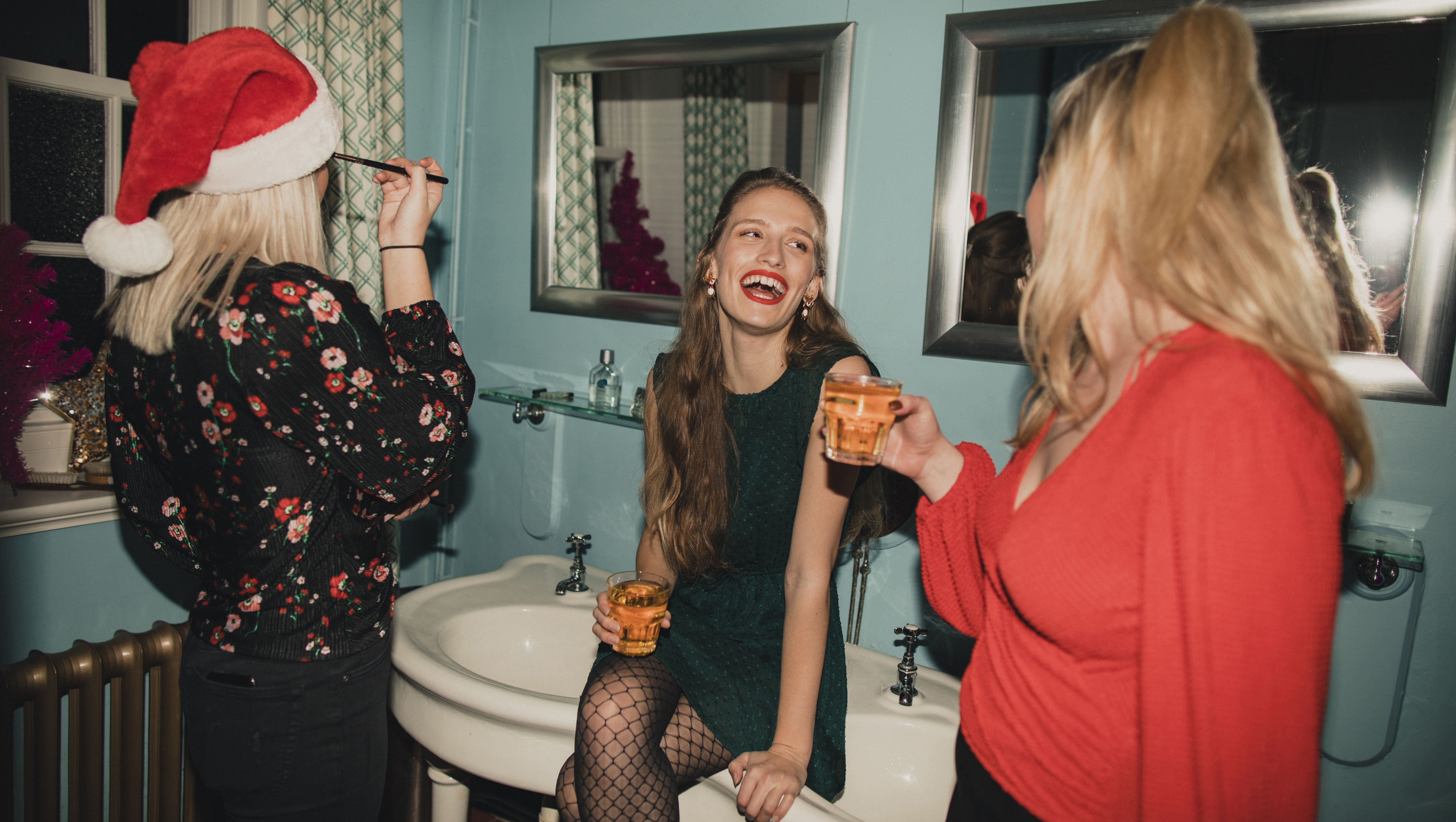 Three young women are drinking and applying makeup in the bathroom at a house party.
