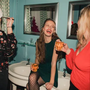 Three young women are drinking and applying makeup in the bathroom at a house party.