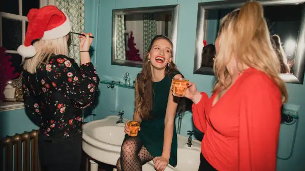 Three young women are drinking and applying makeup in the bathroom at a house party.