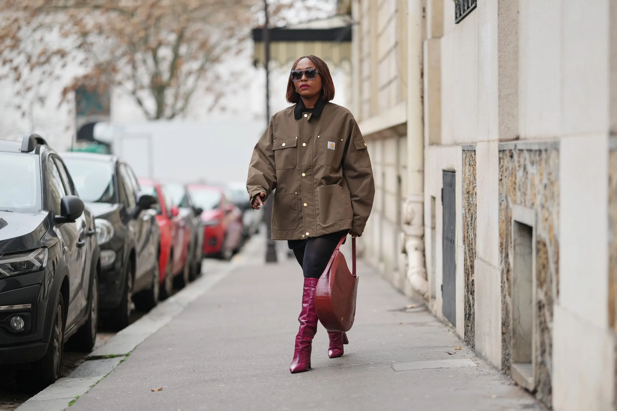 PARIS, FRANCE - DECEMBER 13: Carrole Sagba wears a straight chin-length bob hairstyle with a center part, makeup featuring a natural complexion with soft pink lipstick, an olive green khaki cotton canvas workwear jacket with flap chest pockets and snap-button fastening by Carhartt, black opaque tights, a burgundy leather tote bag with a structured silhouette and crocodile pattern, and burgundy shiny knee-high leather boots with a pointed toe and block heel and crocodile pattern, during a street style fashion photo session, on December 13, 2025 in Paris, France. (Photo by Edward Berthelot/Getty Images)