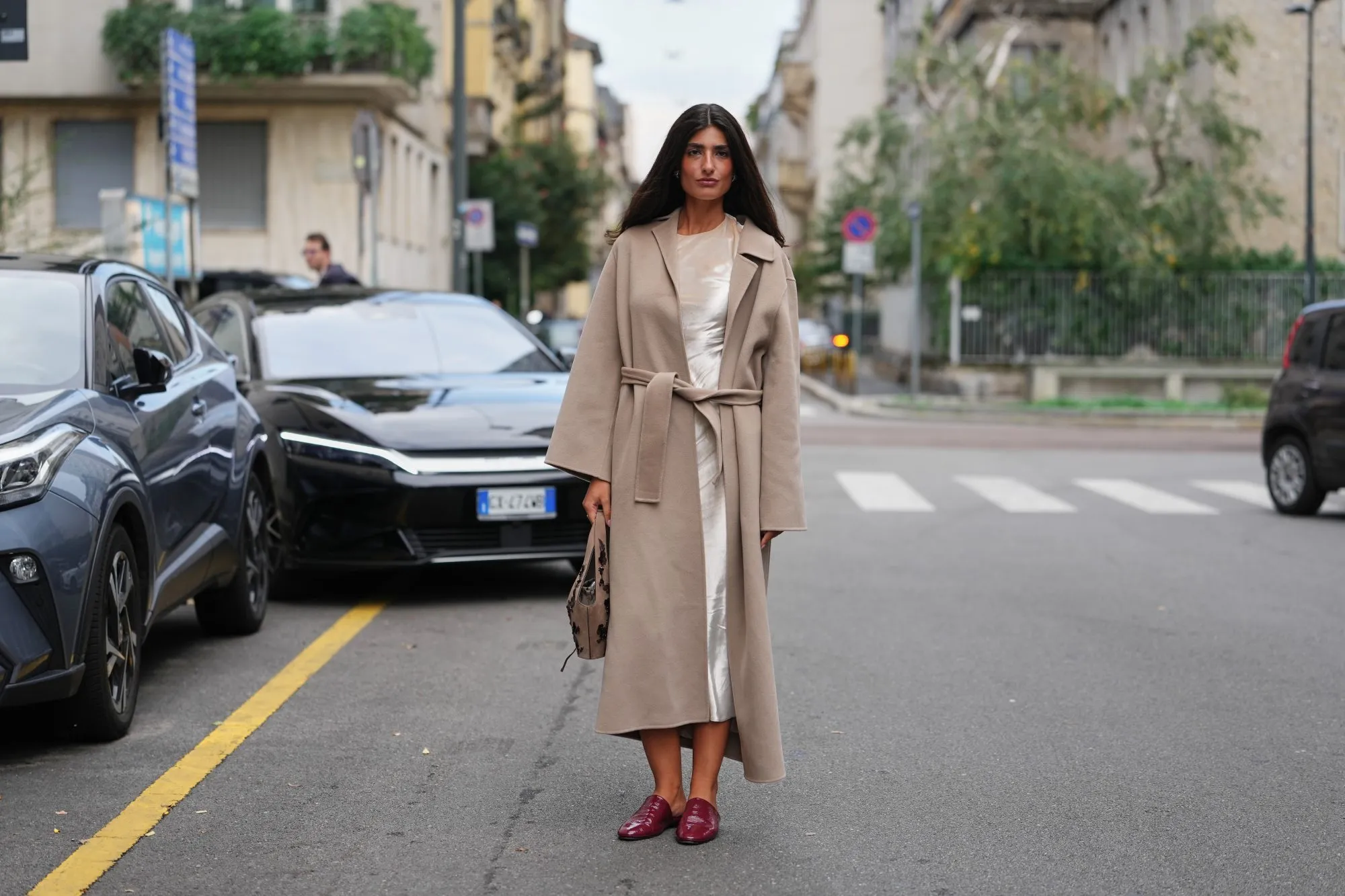 MILAN, ITALY - SEPTEMBER 23: A guest wears long dark brown hair worn loose with a center part, silver stud earrings, a taupe suede hobo bag with dark floral embroidery, a calf-length belted camel-beige wool coat with a wide notched collar and long sleeves, layered over an ivory satin slip dress with visible sheen at the neckline and hem, pointed-toe burgundy patent leather backless mules with a low stacked heel, outside Alberta Ferretti, during Milan Fashion Week - WoWomenswear Spring/Summer 2026, on September 23, 2025 in Milan, Italy (Photo by Edward Berthelot/Getty Images)