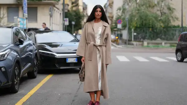 MILAN, ITALY - SEPTEMBER 23: A guest wears long dark brown hair worn loose with a center part, silver stud earrings, a taupe suede hobo bag with dark floral embroidery, a calf-length belted camel-beige wool coat with a wide notched collar and long sleeves, layered over an ivory satin slip dress with visible sheen at the neckline and hem, pointed-toe burgundy patent leather backless mules with a low stacked heel, outside Alberta Ferretti, during Milan Fashion Week - WoWomenswear Spring/Summer 2026, on September 23, 2025 in Milan, Italy (Photo by Edward Berthelot/Getty Images)