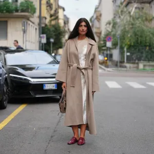 MILAN, ITALY - SEPTEMBER 23: A guest wears long dark brown hair worn loose with a center part, silver stud earrings, a taupe suede hobo bag with dark floral embroidery, a calf-length belted camel-beige wool coat with a wide notched collar and long sleeves, layered over an ivory satin slip dress with visible sheen at the neckline and hem, pointed-toe burgundy patent leather backless mules with a low stacked heel, outside Alberta Ferretti, during Milan Fashion Week - WoWomenswear Spring/Summer 2026, on September 23, 2025 in Milan, Italy (Photo by Edward Berthelot/Getty Images)