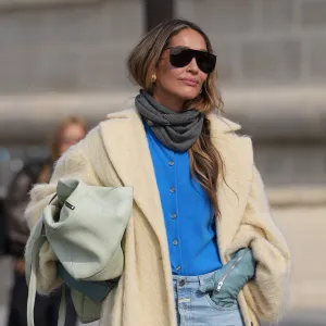 PARIS, FRANCE - OCTOBER 01: A guest wears long wavy light brown hair with a center part, oversized black rectangular sunglasses, small gold earrings, a pale mint green leather puffy shoulder bag, a grey knit scarf, a bright cobalt blue button-up knit cardigan, a long cream fuzzy coat with a light satin lining, pale blue leather gloves, light wash high-rise wide-leg denim jeans, black leather boots shoes with a low heel, outside Dior, during Paris Fashion Week - Womenswear Spring Summer 2026, on October 01, 2025 in Paris, France (Photo by Edward Berthelot/Getty Images)