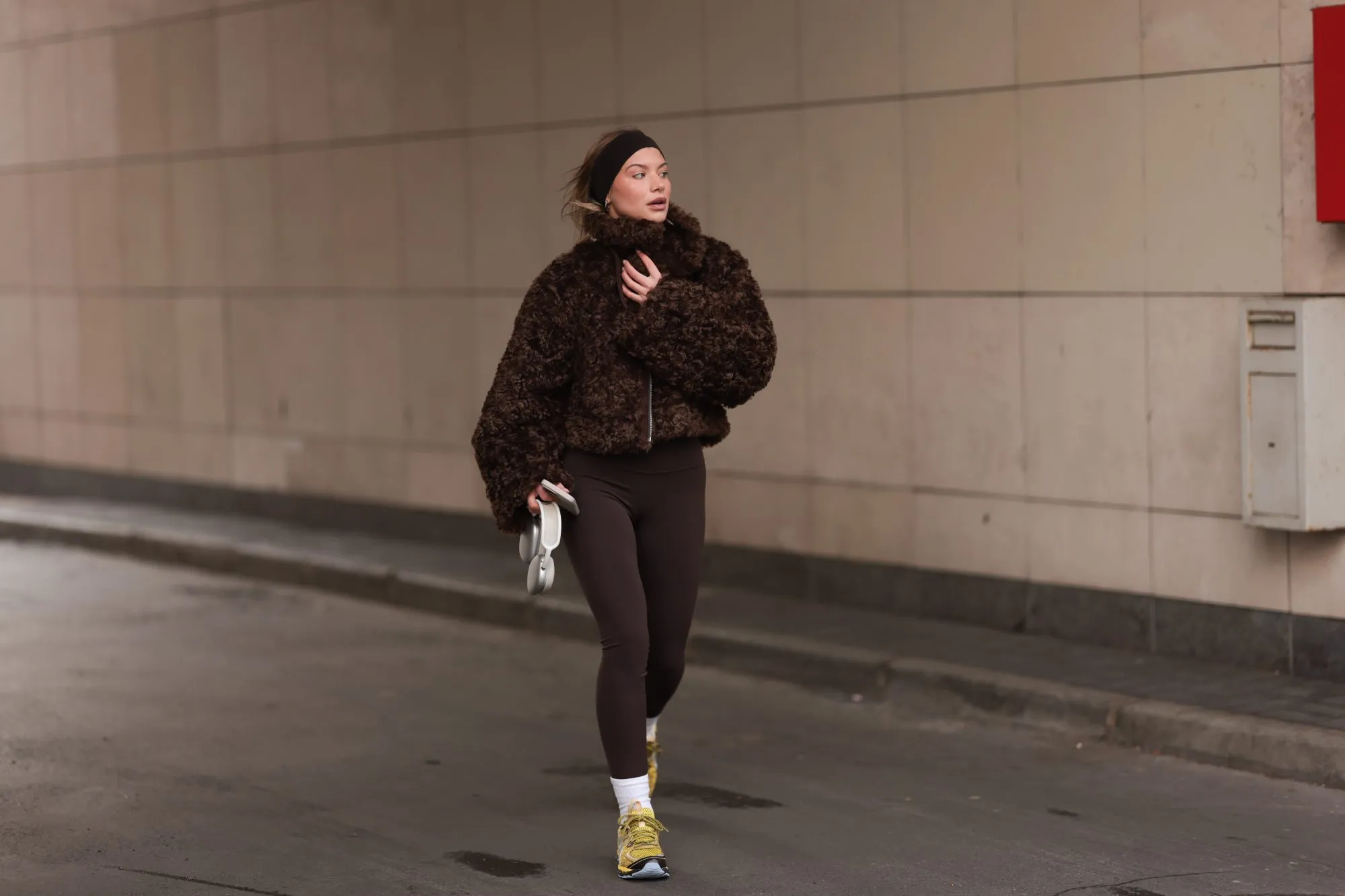 BERLIN, GERMANY - JANUARY 31: Sophia Geiss is seen wearing a dark brown hairband from Naked, two different-sized silver hoop earrings, a silver nose ring, a cropped brown faux shearling jacket with wide sleeves from The Frankie Shop, a dark brown sports bra underneath, paired with dark brown leggings from Lululemon, a silver ring, white socks, and yellow sneakers with light blue highlights and soles from ASICS, while holding AirPods and an iPhone during Berlin Fashion Week on January 31, 2025 in Berlin, Germany. (Photo by Jeremy Moeller/Getty Images)