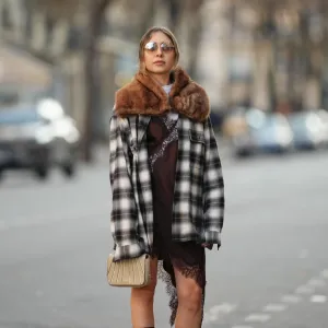 PARIS, FRANCE - DECEMBER 27: Emy Venturini wears light brown hair parted at the center and pulled back, a natural complexion with soft rose-toned lipstick, brown-tinted sunglasses by Prada, a brown faux-fur vintage scarf draped around the shoulders, a black and white plaid long-sleeve shirt by JW Anderson x Uniqlo worn open, layered over a white crew-neck cotton T-shirt by The Frankie Shop, a brown slip-style mini dress by Zara with lace trim visible at the hem, and carries a small beige structured bag by Liase, during a street style fashion photo session, on December 27, 2025 in Paris, France. (Photo by Edward Berthelot/Getty Images)