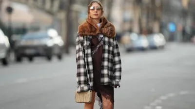 PARIS, FRANCE - DECEMBER 27: Emy Venturini wears light brown hair parted at the center and pulled back, a natural complexion with soft rose-toned lipstick, brown-tinted sunglasses by Prada, a brown faux-fur vintage scarf draped around the shoulders, a black and white plaid long-sleeve shirt by JW Anderson x Uniqlo worn open, layered over a white crew-neck cotton T-shirt by The Frankie Shop, a brown slip-style mini dress by Zara with lace trim visible at the hem, and carries a small beige structured bag by Liase, during a street style fashion photo session, on December 27, 2025 in Paris, France. (Photo by Edward Berthelot/Getty Images)