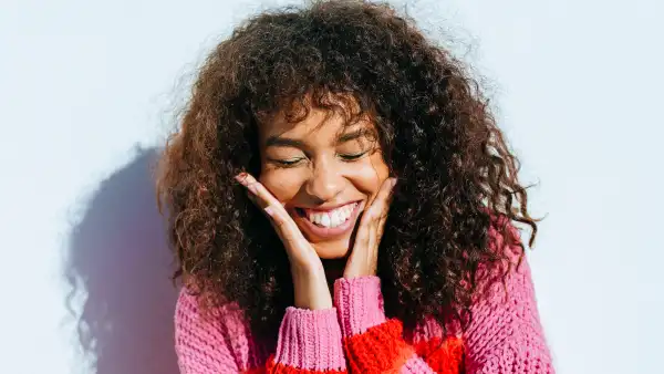Portrait of a cheerful young woman with curly hair wearing colorful sweater, touching her face, smiling and closing her eyes
