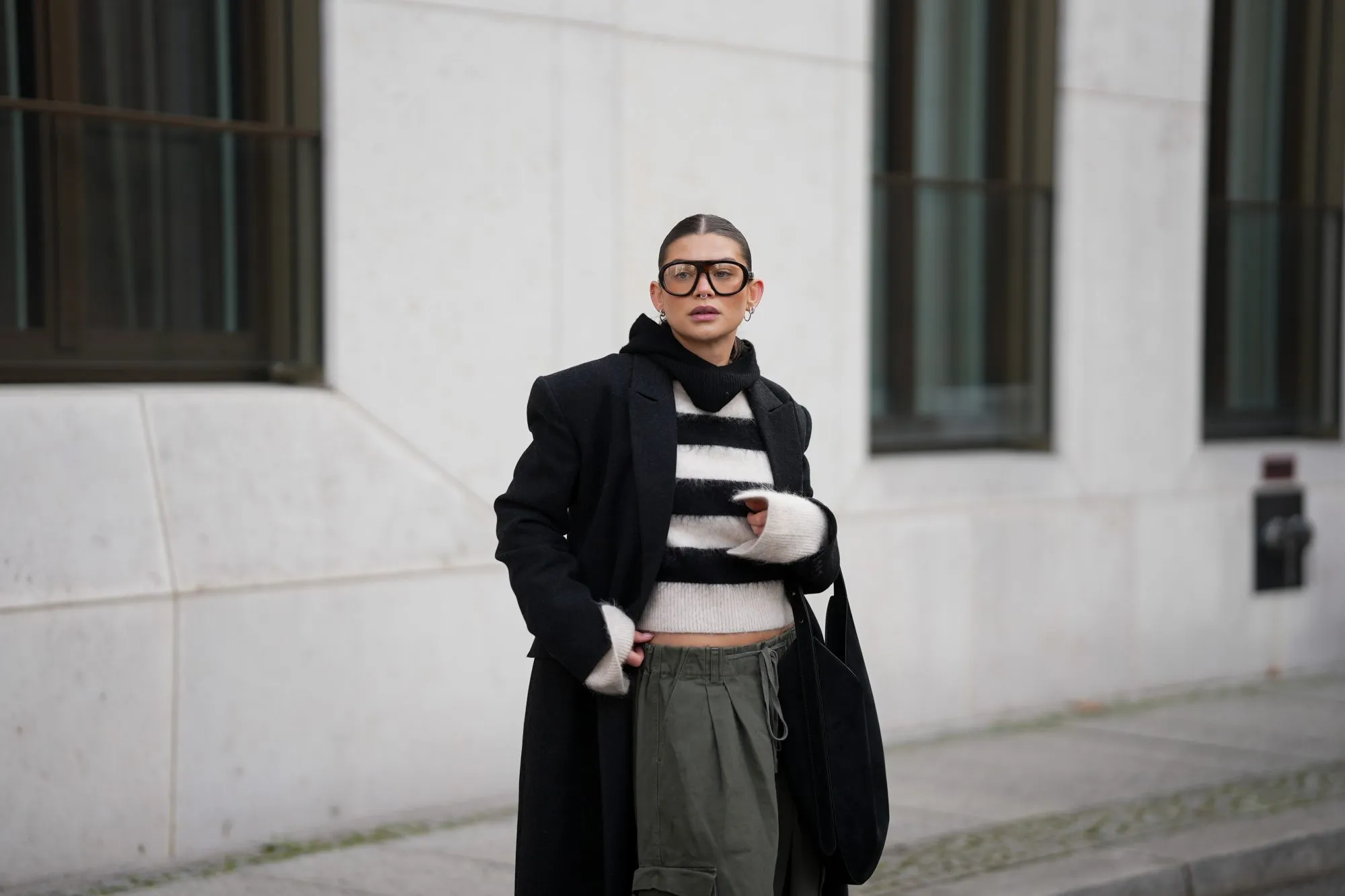 BERLIN, GERMANY - NOVEMBER 25: Sophia Geiss is seen wearing an oversized long black wool coat with structured shoulders and a straight, ankle-length silhouette from Herskind; a slightly cropped, fuzzy black-and-white striped knit sweater from H&M; wide-leg khaki green cargo trousers with drawstring waist and large side pockets from The Frankie Shop; a black suede crescent shoulder hobo bag with a minimalist curved shape from Aesther Ekme; oversized aviator glasses with tortoiseshell frame from Prada; a black knit balaclava from COS; small silver hoop earrings from Maria Black; her brown hair is styled in a sleek low bun with a center parting on November 25, 2025 in Berlin, Germany. (Photo by Moritz Scholz/Getty Images)