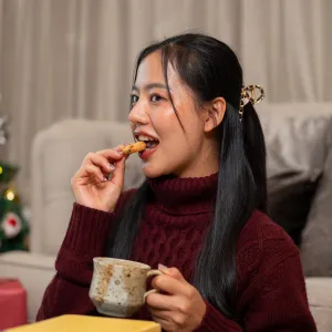 A cheerful, beautiful Asian woman in a cozy red sweater enjoys Christmas cookies and a warm drink while reading in her living room, spending the Christmas holiday peacefully at home alone.