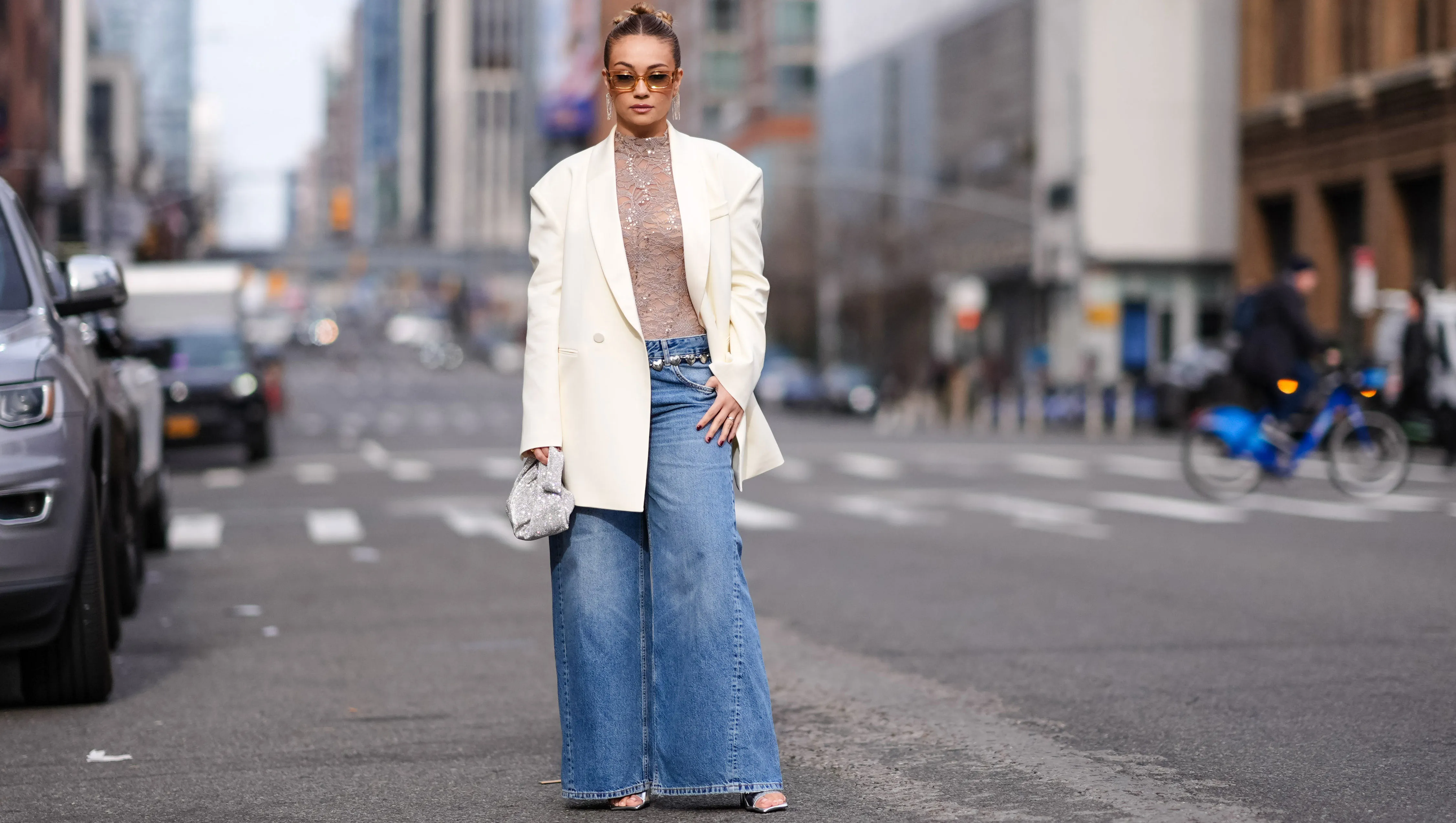 NEW YORK, NEW YORK - FEBRUARY 11: Merve Gorgotz wears sunglasses, earrings, a floral embroidery gray turtleneck top, a white oversized blazer jacket flared blue denim jeans pants , silver shoes, outside Area, during New York Fashion Week, on February 11, 2024 in New York City. (Photo by Edward Berthelot/Getty Images)