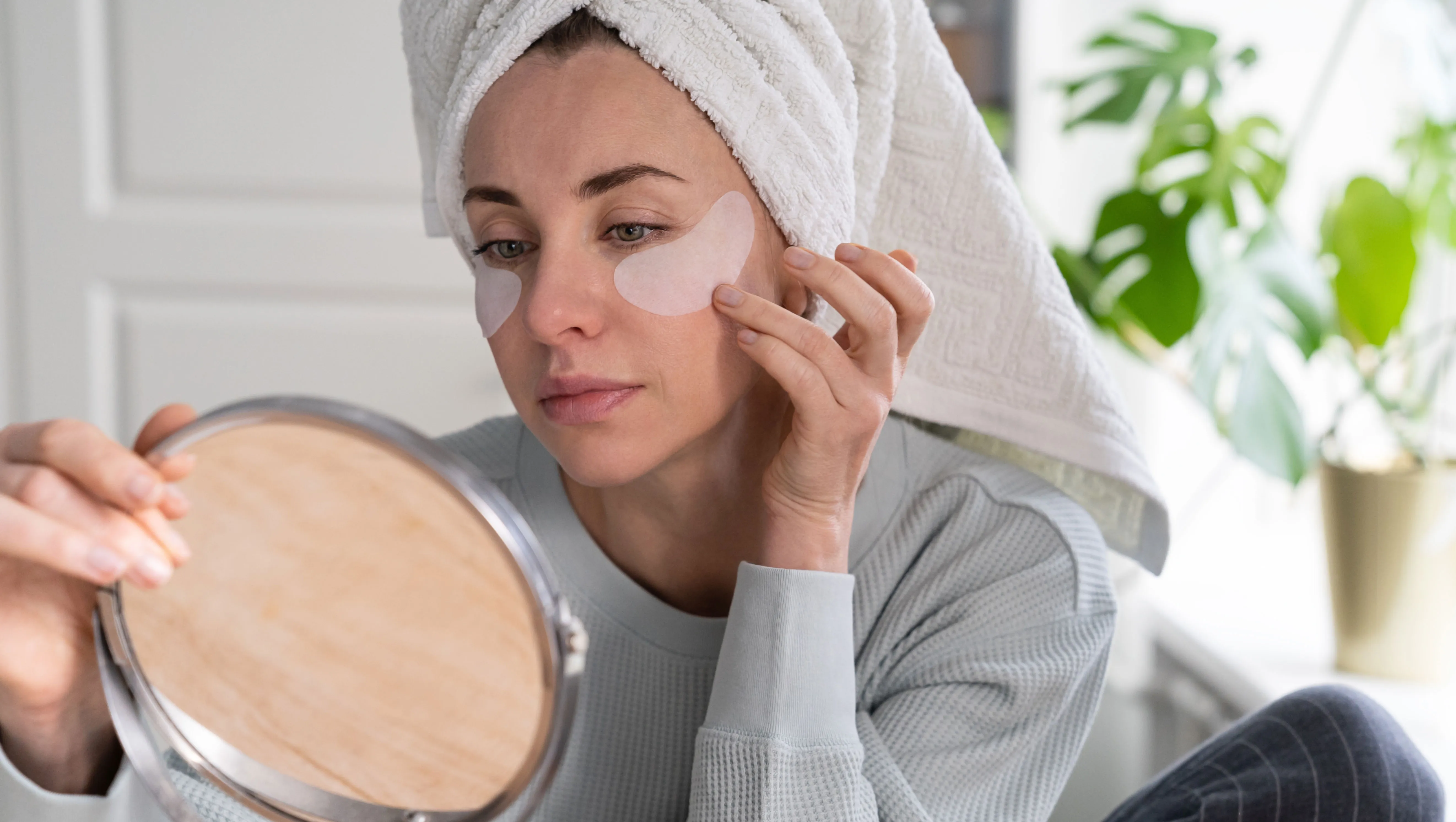 Woman with towel on head applying patches under eyes enriched with collagen, vitamin E, diminishes the signs of aging, helps reduse eye puffiness, looking in mirror. Face skin care beauty at home.