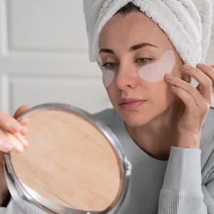 Woman with towel on head applying patches under eyes enriched with collagen, vitamin E, diminishes the signs of aging, helps reduse eye puffiness, looking in mirror. Face skin care beauty at home.