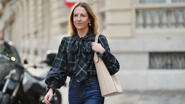 PARIS, FRANCE - DECEMBER 04: Alba Garavito Torre wears long brown hair parted at the center with natural skin and neutral lipstick, a long-sleeve plaid shirt in black green and blue with an oversized neck bow by Frambua, dark-indigo high-rise flare jeans pants by Citizens of Humanity, a beige smooth calf-leather hobo bag with a curved design by Studio Flo, gold earrings and a gold ring by Sempiterno Paris, during a street style fashion photo session, on December 04, 2025 in Paris, France, full body shot. (Photo by Edward Berthelot/Getty Images)
