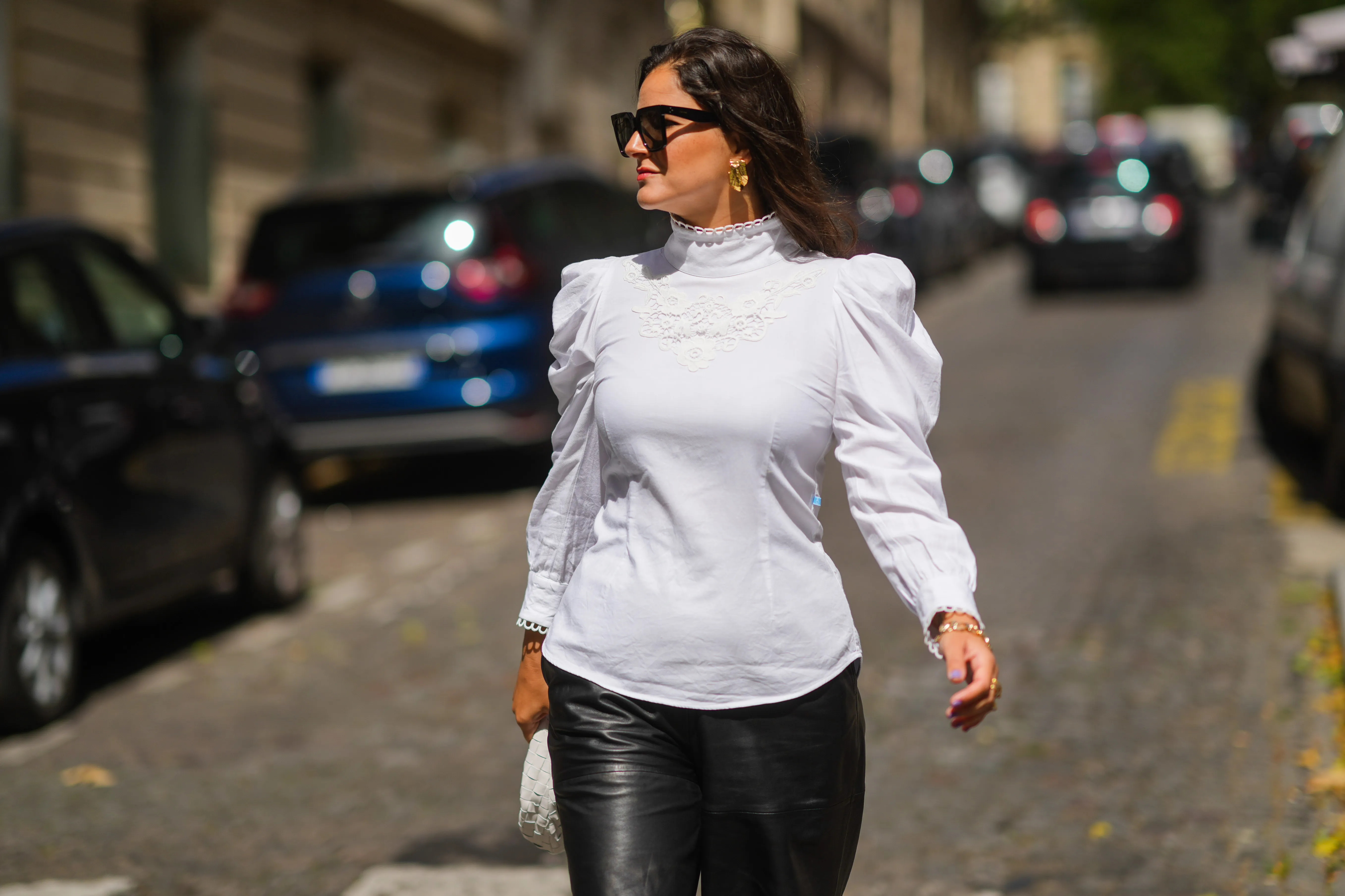 Gili Biegun wears black sunglasses, gold earrings, a white turtleneck puffy shoulder long sleeves shirt with embroidered, black shiny leather shorts, a large chain gold bracelet, a white braided leather handbag from Bottega Veneta, outside Chanel, during Paris Fashion Week - Haute Couture Fall/Winter 2021/2022, on July 06, 2021 in Paris, France.