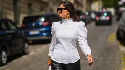 Gili Biegun wears black sunglasses, gold earrings, a white turtleneck puffy shoulder long sleeves shirt with embroidered, black shiny leather shorts, a large chain gold bracelet, a white braided leather handbag from Bottega Veneta, outside Chanel, during Paris Fashion Week - Haute Couture Fall/Winter 2021/2022, on July 06, 2021 in Paris, France.