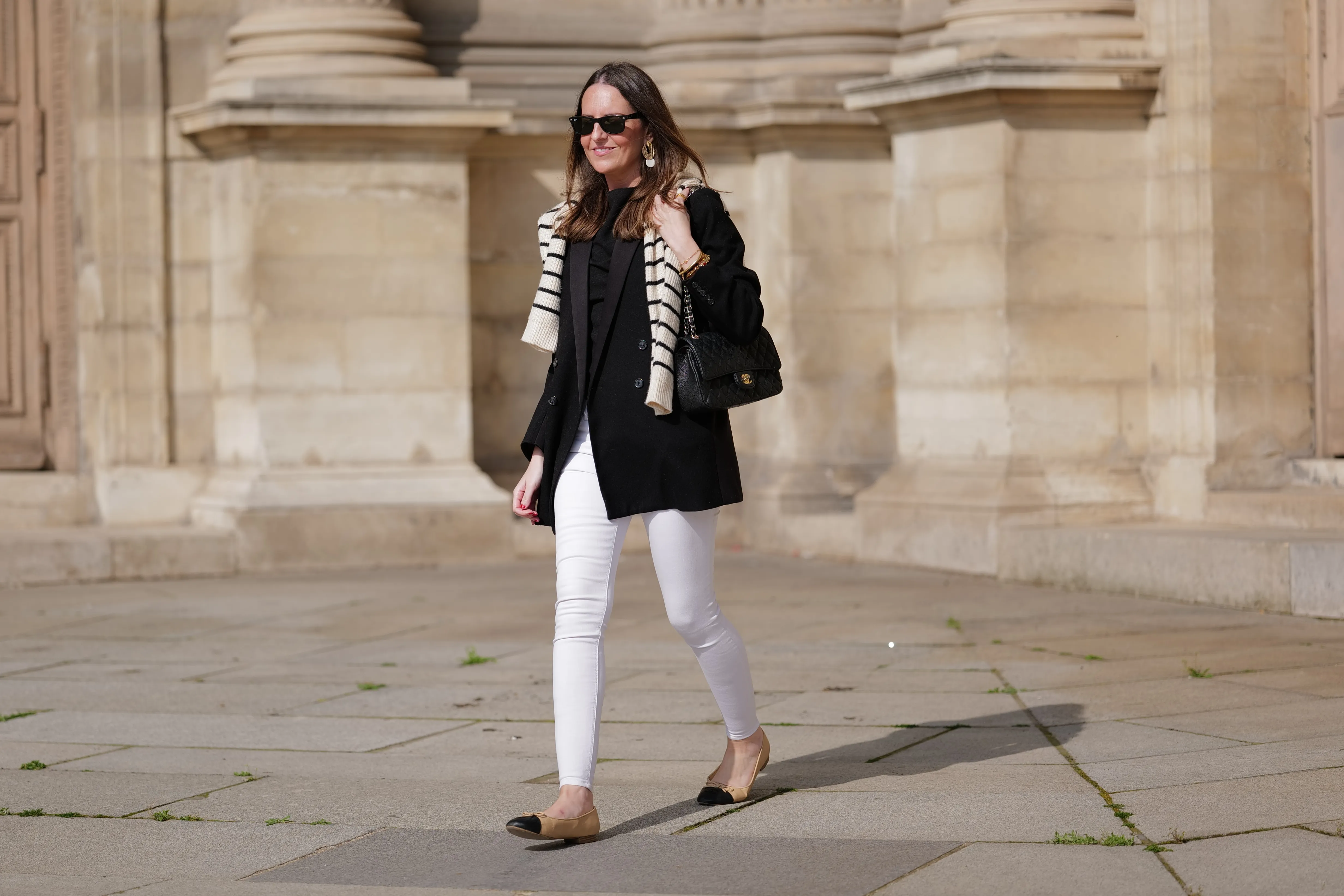 Alba Garavito Torre wears black sunglasses from Ray Ban, gold and white pendant large earrings, a black t-shirt, a white with black small striped print pattern wool pullover from Massimo Dutti, a black shiny grained leather Timeless shoulder bag from Chanel, a black oversized blazer jacket from Munthe, white skinny denim pants from Zara, beige shiny leather with black toe-cap ballerinas from Chanel , during a street style fashion photo session, on April 29, 2023 in Paris, France.