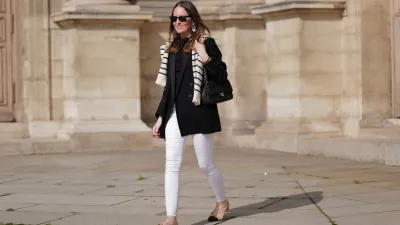 Alba Garavito Torre wears black sunglasses from Ray Ban, gold and white pendant large earrings, a black t-shirt, a white with black small striped print pattern wool pullover from Massimo Dutti, a black shiny grained leather Timeless shoulder bag from Chanel, a black oversized blazer jacket from Munthe, white skinny denim pants from Zara, beige shiny leather with black toe-cap ballerinas from Chanel , during a street style fashion photo session, on April 29, 2023 in Paris, France.