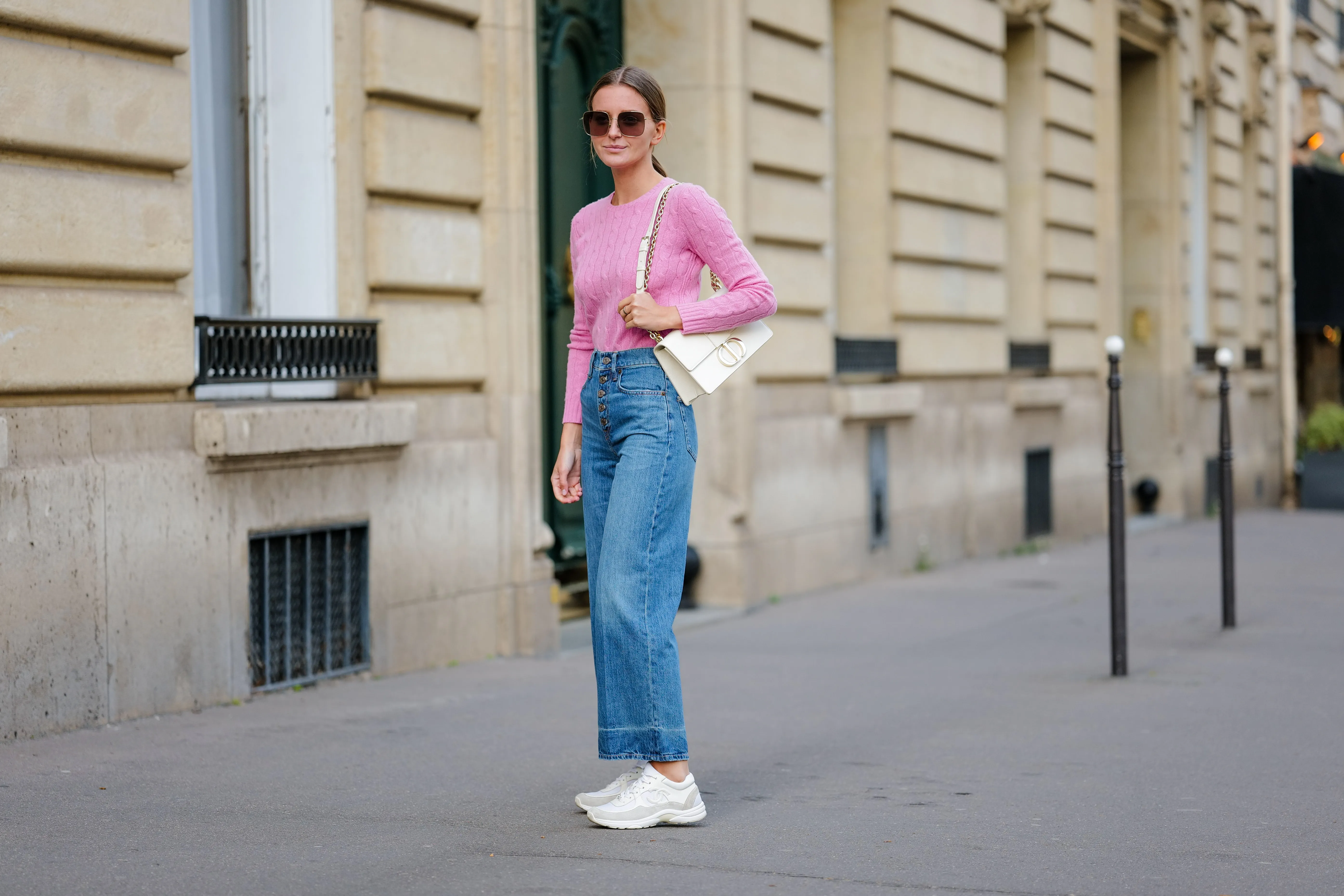 Diane Batoukina wears dark brown large squared sunglasses from Dior, a pale pink braided wool pullover from Ralph Lauren, blue faded denim / buttons high waist / wide legs pants, a white latte matte leather 30 Montaigne handbag from Dior, white suede and leather sneakers from Chanel, during a street style fashion photo session, on April 21, 2023 in Paris, France.