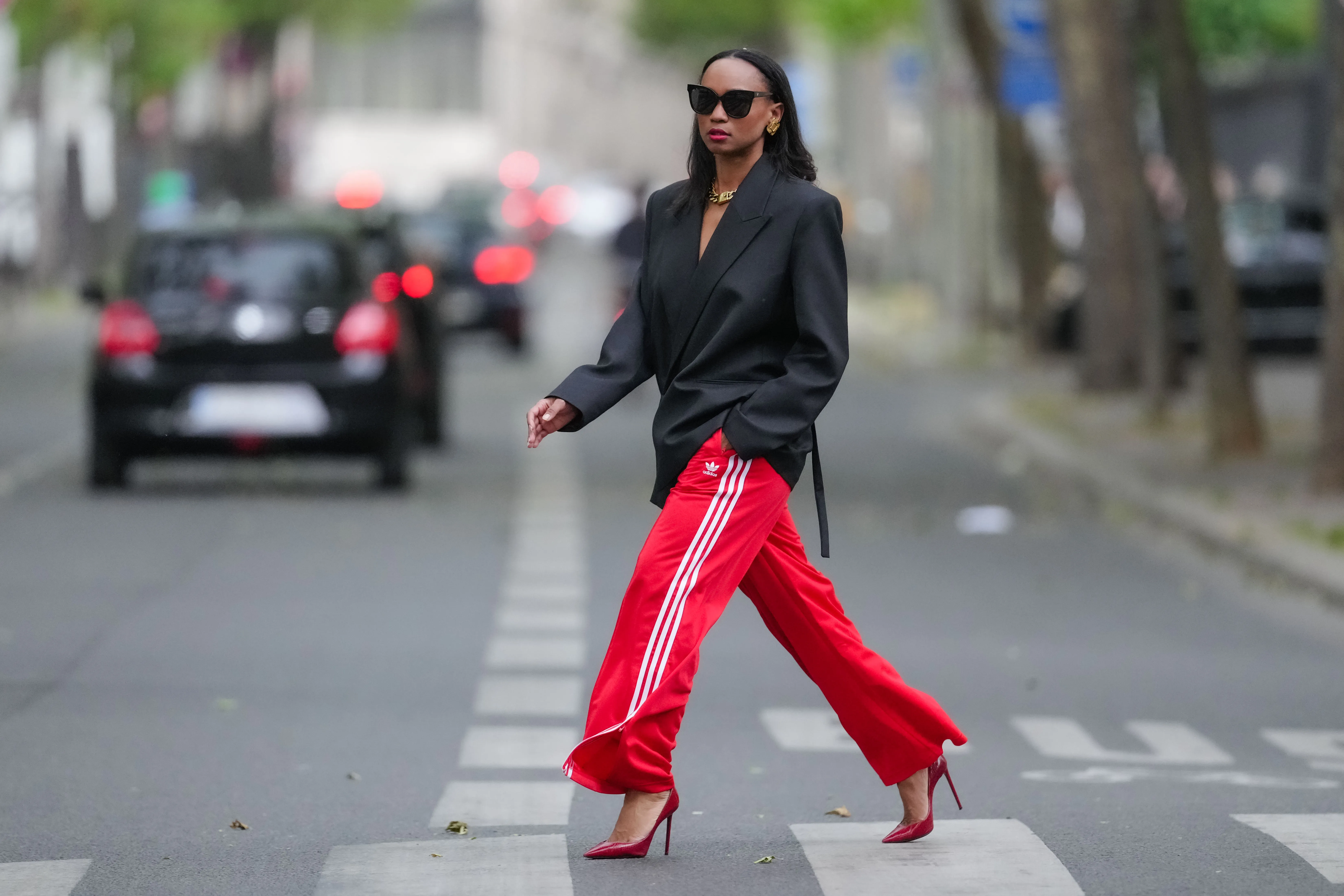 Emilie Joseph wears sunglasses, earrings, red lipstick, a black oversized / structured virgin wool blend Blazer with lapel-collar by Victoria Beckham for Mango, Adidas Classics oversized tracksuit bottoms / track pants in red with stripes, a golden chunky necklace / chain by Merbabe Greece, shoes : Newton satin pointy black pumps with high heels by Saint Laurent, during a street style fashion photo session, on May 12, 2024 in Paris, France.