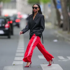 Emilie Joseph wears sunglasses, earrings, red lipstick, a black oversized / structured virgin wool blend Blazer with lapel-collar by Victoria Beckham for Mango, Adidas Classics oversized tracksuit bottoms / track pants in red with stripes, a golden chunky necklace / chain by Merbabe Greece, shoes : Newton satin pointy black pumps with high heels by Saint Laurent, during a street style fashion photo session, on May 12, 2024 in Paris, France.