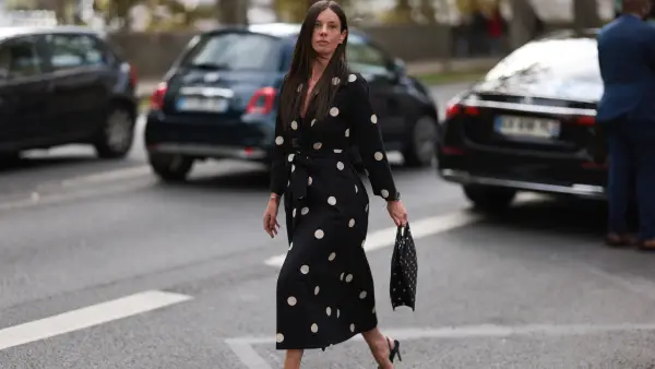 A Fashion Week guest is seen wearing round diamond stud earrings, a long-sleeved black midi dress with white polka dot pattern, notched lapels, and a tie belt; a gold bangle; a rose gold and black watch from Rolex; a gold ring; a rectangular black handbag with a white polka dot pattern; and pointed black slingback pumps, also with a white polka dot pattern before the Elie Saab Womenswear Spring/Summer 2025 Show as part of Paris Fashion Week on September 28, 2024 in Paris, France.