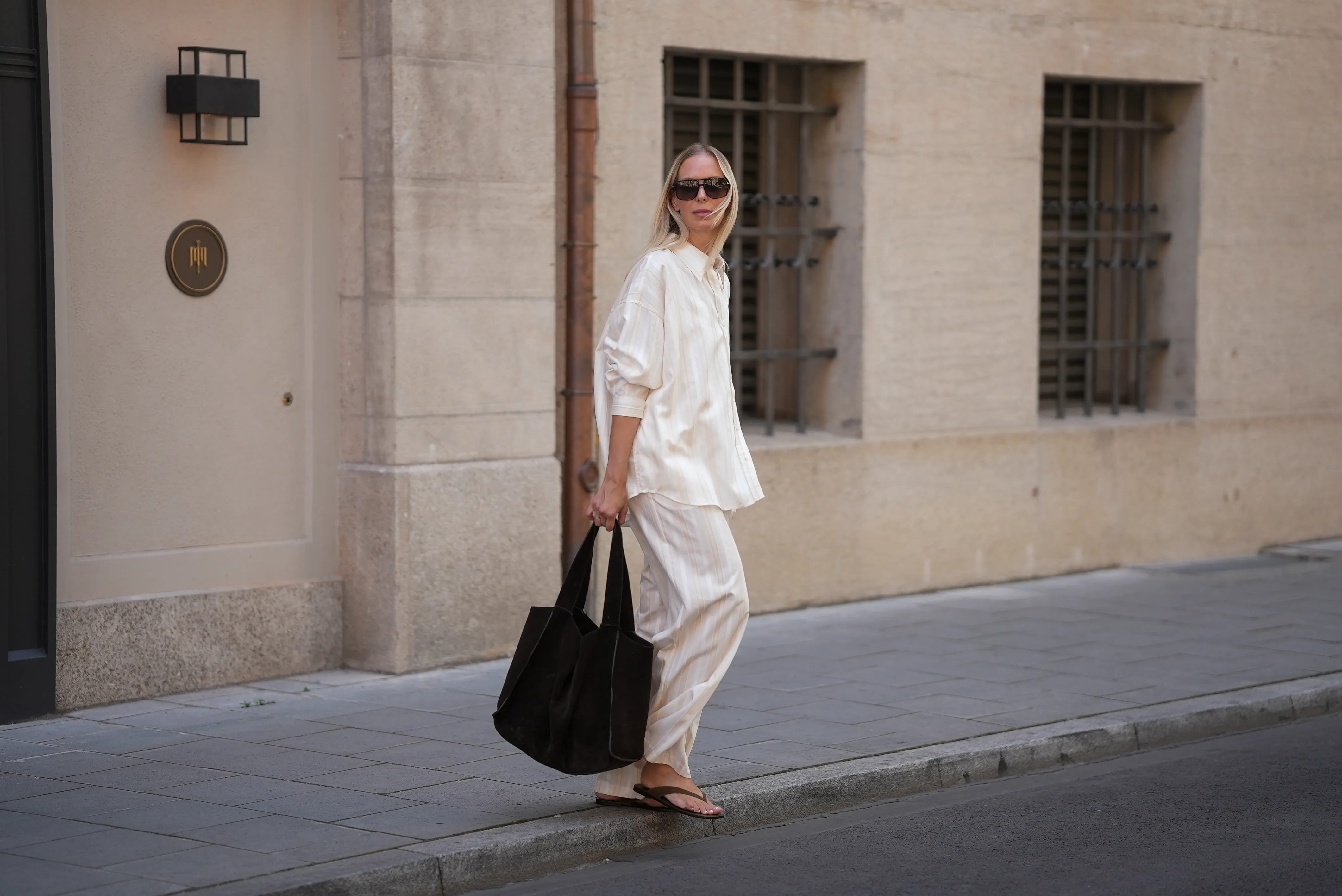 Mirja Klein is seen wearing a matching set consisting of a white striped oversized dobby-weave shirt with collar and buttons down in the front from H&M and a white striped straight pull-on trousers with loose fit and straight legs from H&M; a black suede tote bag with slouchy design and open top from COS; black oversized aviator sunglasses from Tom Ford; a pair of khaki green suede flip flops from A.EMERY on June 18, 2025 in Munich, Germany.