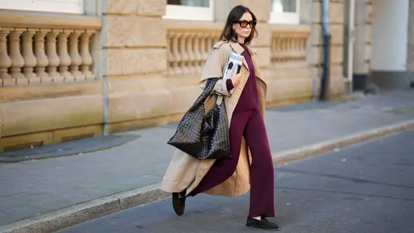 Patricia Wirschke is seen wearing a classic beige long Herno trench coat, with a notched collar, storm flap, and belted cuffs; a burgundy Incentive co ord set with a cable-knit sweater, featuring a ribbed hem, cuffs, and neckline and wide-leg knit pants with a relaxed fit; a pair of Le Loafer soft suede Saint Laurent moccasins in dark brown with golden logo detail on the side; a dark brown Hop bag from Bottega Veneta with the famous Intrecciato leather construction, woven; Tom Ford Ilias squared frame black sunglasses with orange toned lens; Bvlgari Serpenti Viper golden earrings; her brown hair is styled in a straight, shoulder-length look with a middle part; she holds a rolled up magazine in her hand on August 19, 2025 in Duesseldorf, Germany.