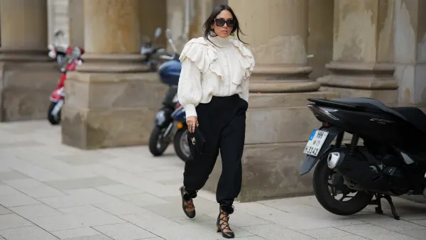 Jennifer Casimiro is seen wearing a cream cable-knit turtleneck sweater with voluminous ruffled detailing on the shoulders from Zara; black high-waisted trousers with a tapered silhouette, cinched at the ankles from Zara; black lace-up shoes with block heels, featuring pom-pom accents at the ankle ties from Gucci; the Suede Varenne small black clutch bag with a rectangular shape and embossed gold branding from Jimmy Choo; oversized black rectangular sunglasses from MiuMiu; gold cuffed earrings from Zara; her black hair is worn down with a center part on September 19, 2025 in Hamburg, Germany.