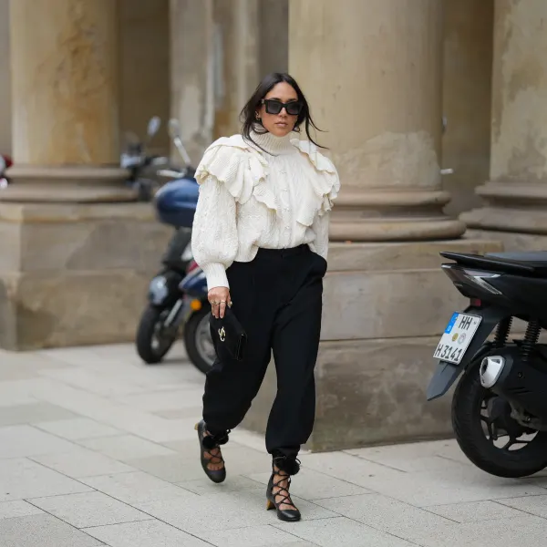 Jennifer Casimiro is seen wearing a cream cable-knit turtleneck sweater with voluminous ruffled detailing on the shoulders from Zara; black high-waisted trousers with a tapered silhouette, cinched at the ankles from Zara; black lace-up shoes with block heels, featuring pom-pom accents at the ankle ties from Gucci; the Suede Varenne small black clutch bag with a rectangular shape and embossed gold branding from Jimmy Choo; oversized black rectangular sunglasses from MiuMiu; gold cuffed earrings from Zara; her black hair is worn down with a center part on September 19, 2025 in Hamburg, Germany.