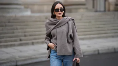 Laura Noltemeyer is seen wearing a taupe fine-knit sweater with a sculptural draped front collar from Zara; light-blue straight-leg denim jeans with a soft fading from Zara; beige suede winter boots with a rounded toe, rope laces, tonal stitching and a thick rubber sole from Inuikii;a large textured dark-brown shoulder bag in a soft sculptural, slouchy shape and woven leather from Les Visionnaires x Michi from the Want; black oval sunglasses with a glossy acetate frame and dark lenses from Celine; chunky gold sculptural earrings from Zara; dark brown soft leather gloves from Zara; her dark brown hair is worn straight with a middle parting, tucked behind the ears on November 25, 2025 in Berlin, Germany.