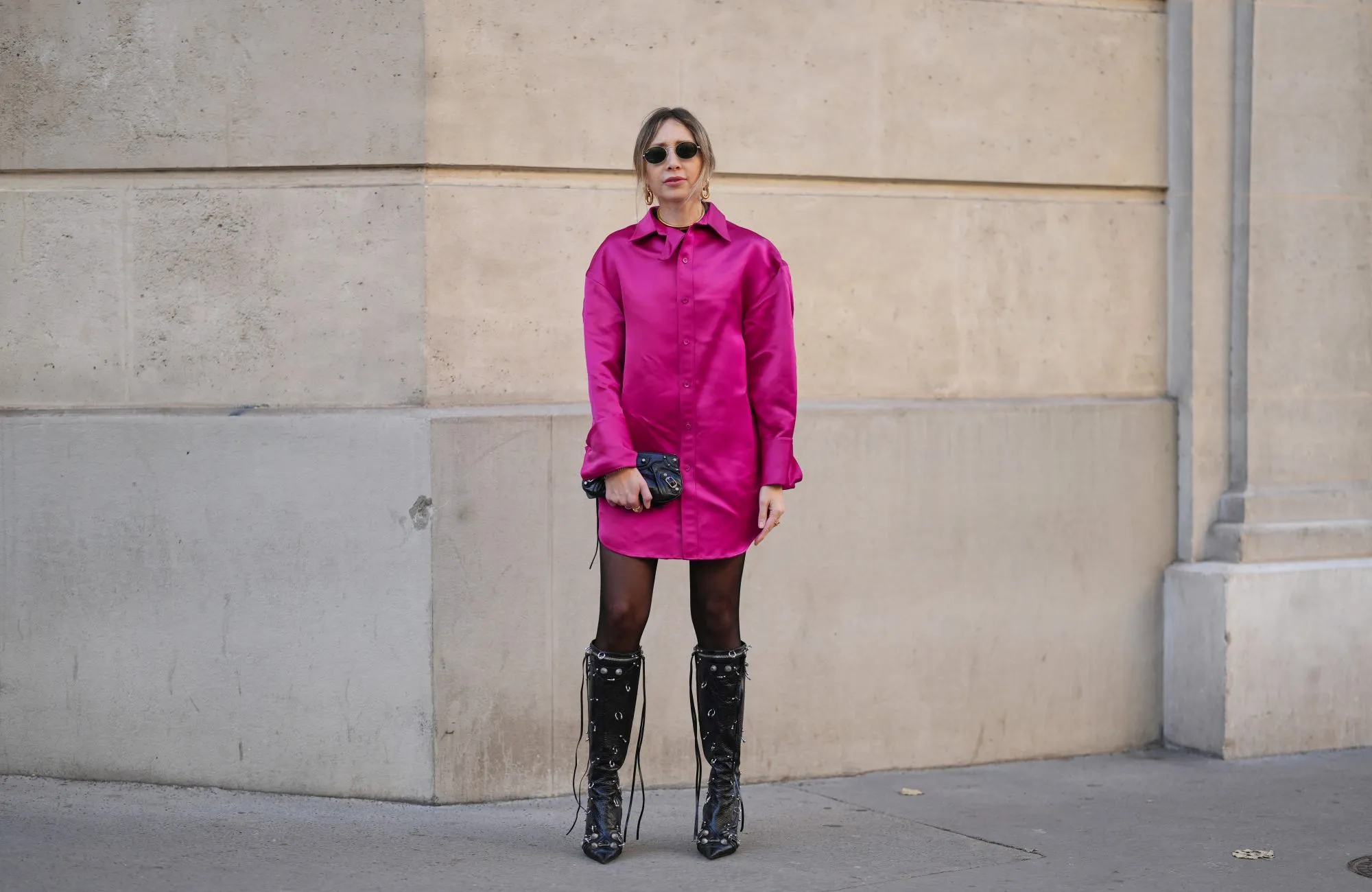 PARIS, FRANCE - DECEMBER 05: Emy Venturini wears light brown hair tied back with loose strands, natural skin with neutral lipstick, small oval sunglasses by Ray Ban, a bright fuchsia neon pink satin long sleeve shirt dress by Balenciaga, sheer black tights by Calzedonia, black leather lace up knee high boots shoes with metal eyelet details by Balenciaga, a black leather Balenciaga clutch bag with visible hardware, a vintage gold tone YSL necklace worn close to the neck, during a street style fashion photo session, on December 05, 2025 in Paris, France. (Photo by Edward Berthelot/Getty Images)