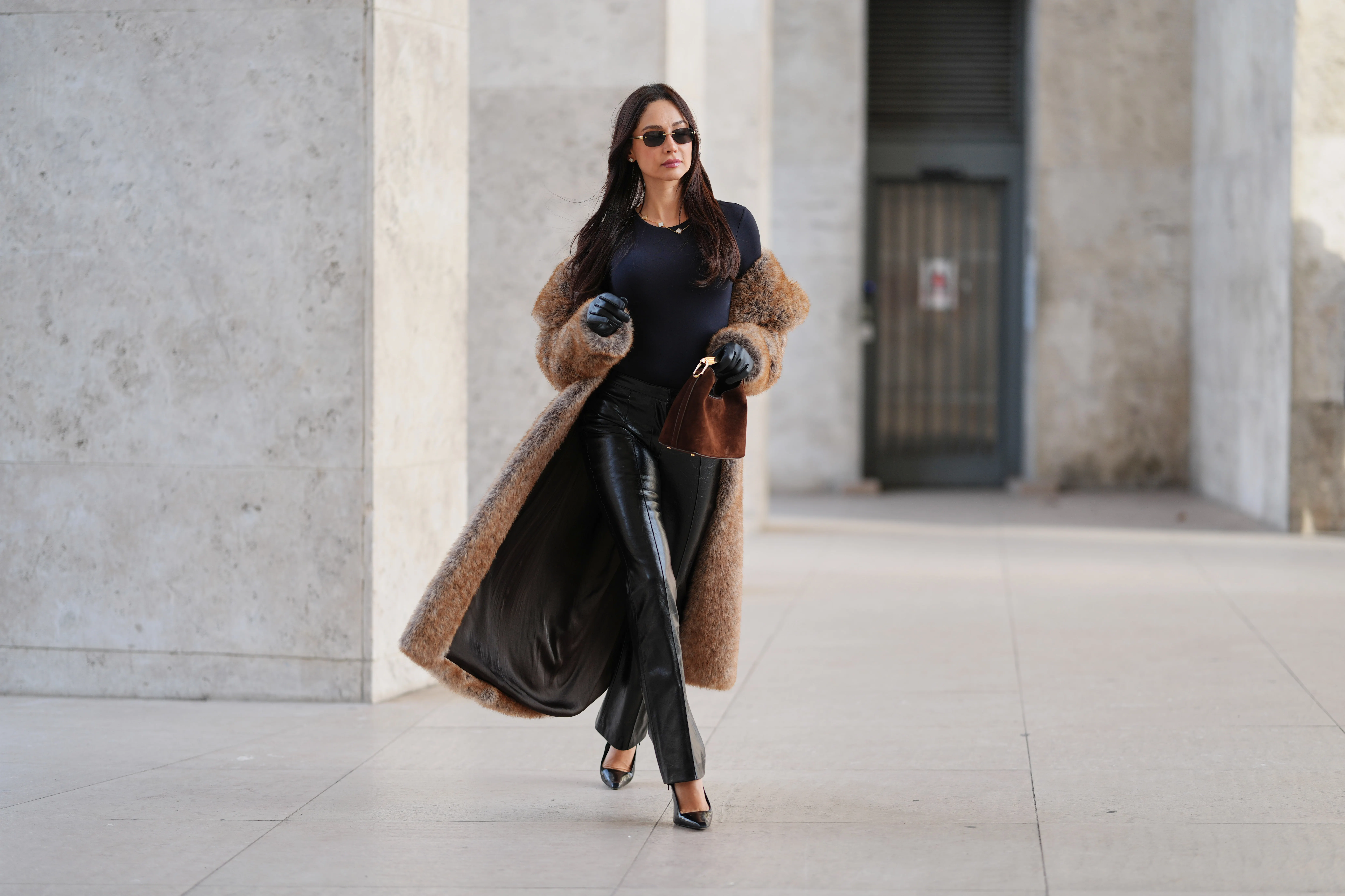 Patricia Gloria Contreras wears long straight dark brown hair worn loose, natural skin with neutral lipstick, narrow rectangular black sunglasses, a long brown faux fur coat by The Frankie Shop worn open, a black fitted long sleeve top with a round neckline, black high waisted straight leg leather pants by Mango, black leather gloves, black pointed toe high heel pumps by Saint Laurent, and a brown suede mini handbag with a curved top handle by Elleme, during a street style fashion photo session, on December 09, 2025 in Paris, France.