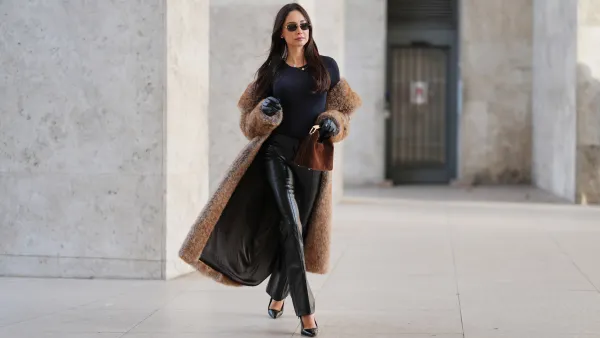 Patricia Gloria Contreras wears long straight dark brown hair worn loose, natural skin with neutral lipstick, narrow rectangular black sunglasses, a long brown faux fur coat by The Frankie Shop worn open, a black fitted long sleeve top with a round neckline, black high waisted straight leg leather pants by Mango, black leather gloves, black pointed toe high heel pumps by Saint Laurent, and a brown suede mini handbag with a curved top handle by Elleme, during a street style fashion photo session, on December 09, 2025 in Paris, France.