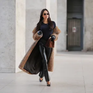 Patricia Gloria Contreras wears long straight dark brown hair worn loose, natural skin with neutral lipstick, narrow rectangular black sunglasses, a long brown faux fur coat by The Frankie Shop worn open, a black fitted long sleeve top with a round neckline, black high waisted straight leg leather pants by Mango, black leather gloves, black pointed toe high heel pumps by Saint Laurent, and a brown suede mini handbag with a curved top handle by Elleme, during a street style fashion photo session, on December 09, 2025 in Paris, France.