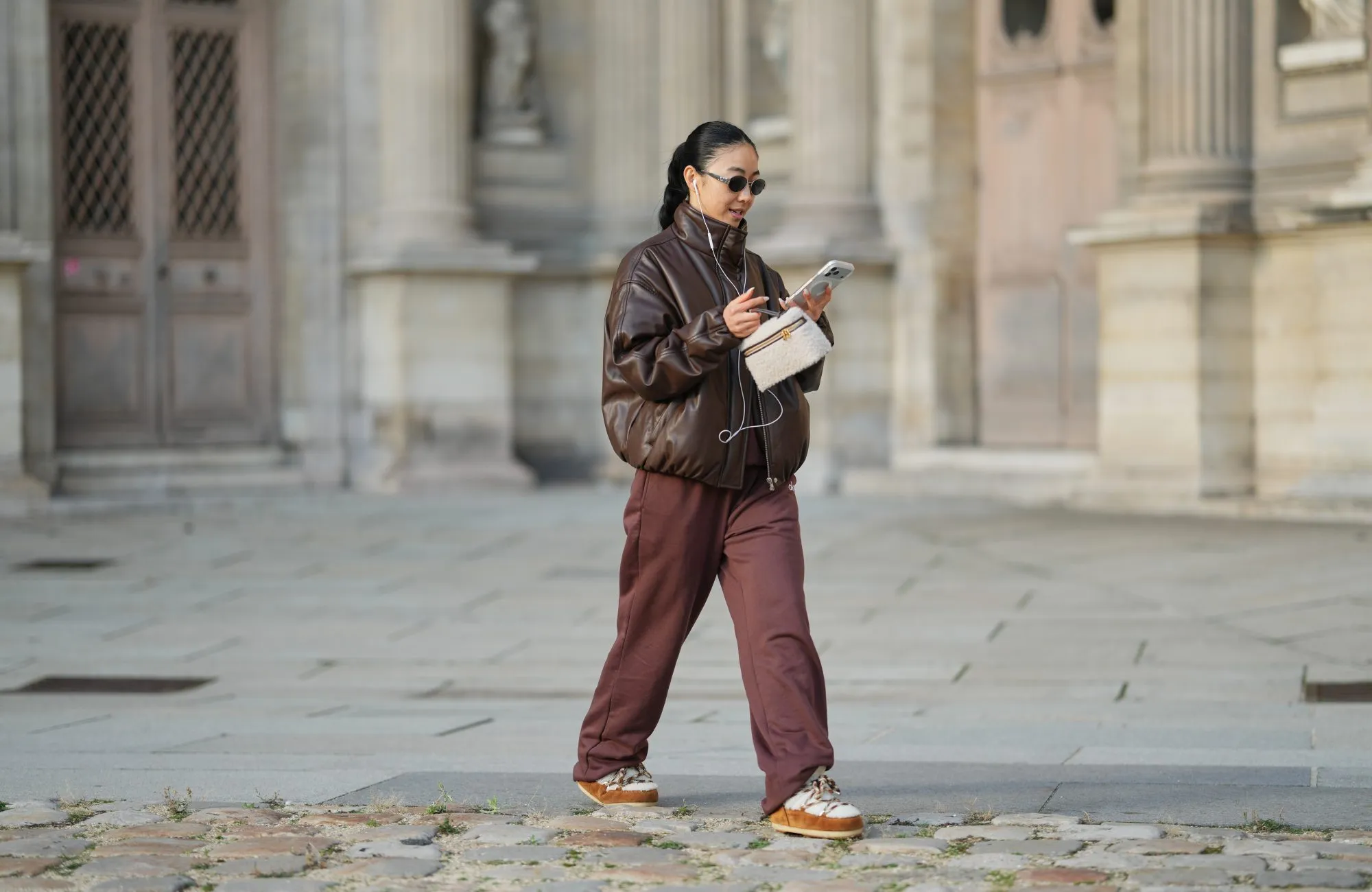 PARIS, FRANCE - DECEMBER 31: Yuwei Zhangzou wears hair pulled back into a low braided ponytail, makeup with an even complexion, defined brows and lipstick, wire earphones, oval dark-lens silver sunglasses by Celine, a brown oversized bomber leather jacket with a high collar and front zip closure, straight-leg brown sport jogger pants by Alo, a small structured beige and brown shearling fluffy bag with a top handle by Loro Piana, white shearling-lined lace-up shoes with brown suede panels, contrast laces and a flat rubber sole by Moon Boot, holds a mobile phone, during a street style fashion photo session, on December 31, 2025 in Paris, France. (Photo by Edward Berthelot/Getty Images)