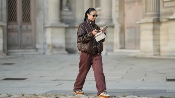 PARIS, FRANCE - DECEMBER 31: Yuwei Zhangzou wears hair pulled back into a low braided ponytail, makeup with an even complexion, defined brows and lipstick, wire earphones, oval dark-lens silver sunglasses by Celine, a brown oversized bomber leather jacket with a high collar and front zip closure, straight-leg brown sport jogger pants by Alo, a small structured beige and brown shearling fluffy bag with a top handle by Loro Piana, white shearling-lined lace-up shoes with brown suede panels, contrast laces and a flat rubber sole by Moon Boot, holds a mobile phone, during a street style fashion photo session, on December 31, 2025 in Paris, France. (Photo by Edward Berthelot/Getty Images)